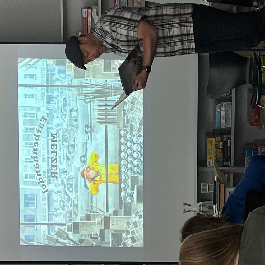 Ein Mann in einem karierten Hemd und einer Kappe steht vor einer Projektionsleinwand, hält ein Buch und einen Stift. Die Leinwand zeigt ein Buchcover mit dem Titel 'Erpeguenagel'. Schüler sitzen darunter.