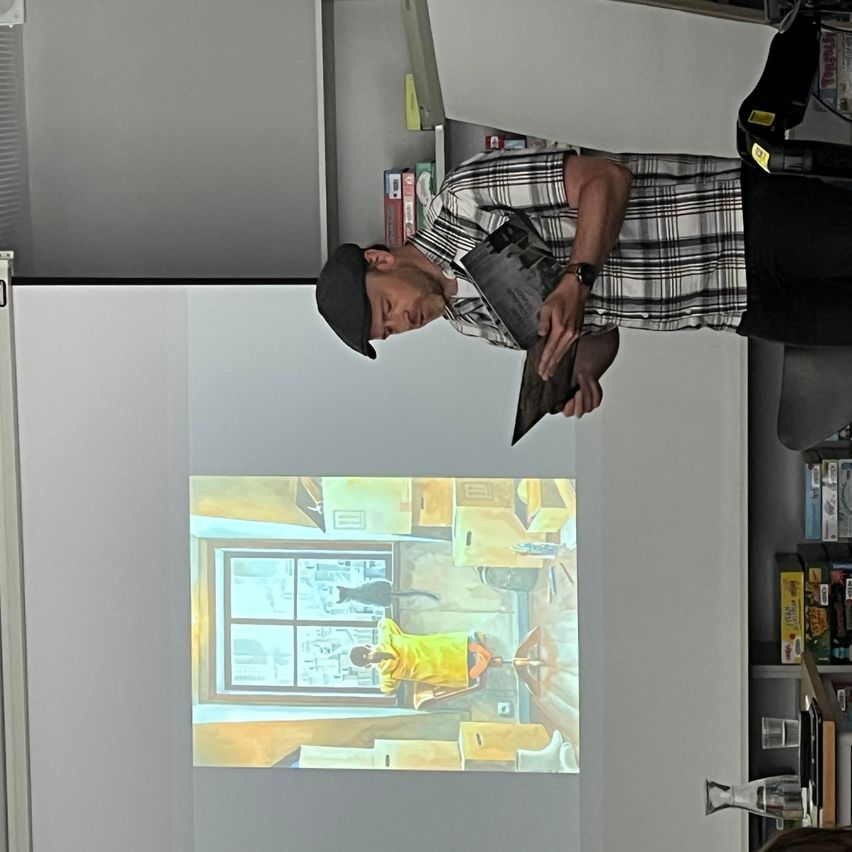 Ein Mann steht vor einer Leinwand, die ein Gemälde zeigt. Er trägt ein kariertes Hemd und einen Hut. Das Gemälde zeigt eine Person in einem gelben Shirt.