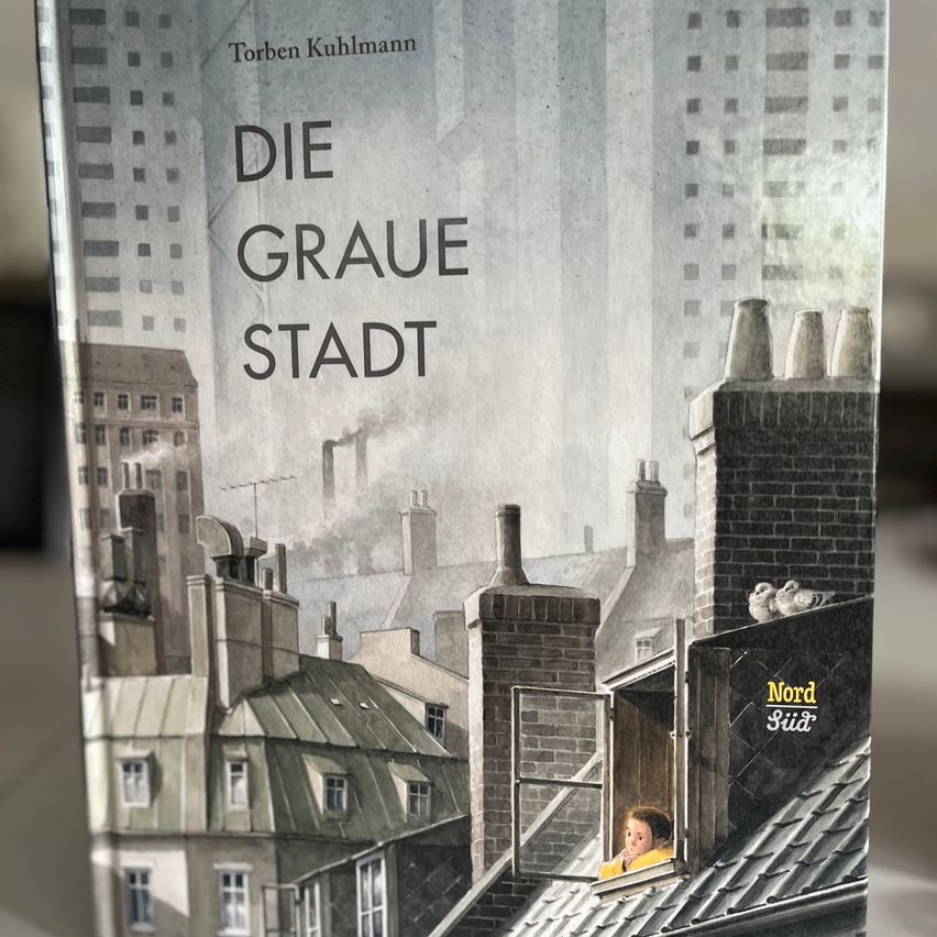 Ein Buchcover mit einer monochromen Stadtlandschaft, die ein Kind in einem Fenster zeigt. Das Buch heißt 'Die Graue Stadt' und es hat den Namen des Autors 'Torben Kuhlmann' darauf.
