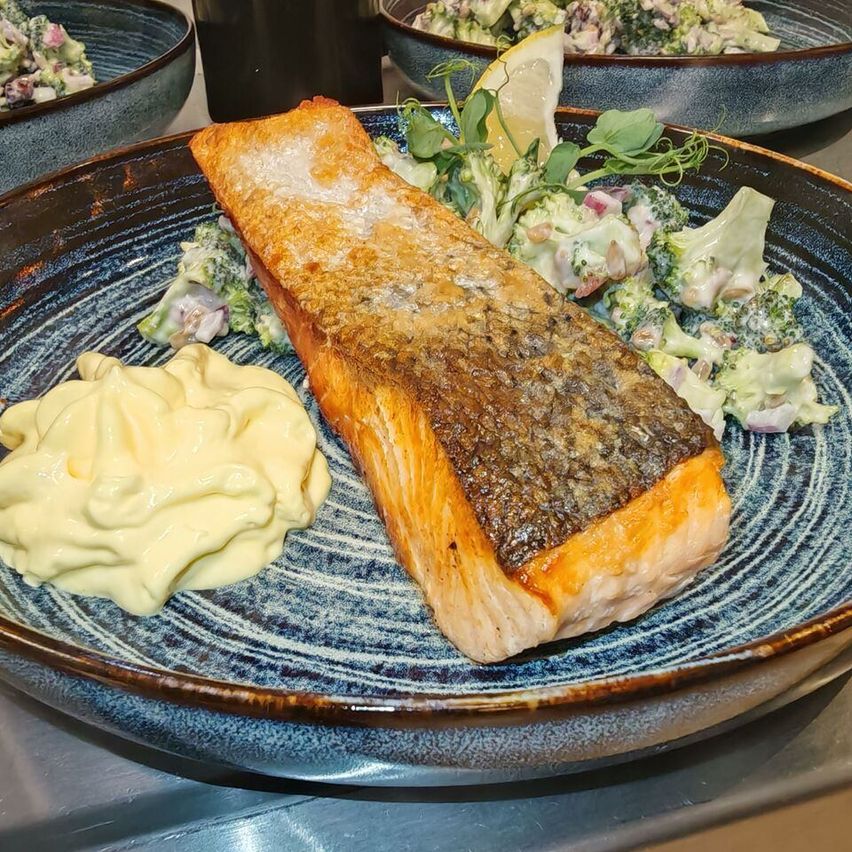 Ein blauer Teller mit einem gegrillten Lachsfilet, Brokkoli-Salat und einer cremigen Dip. Andere Gerichte sind im Hintergrund.