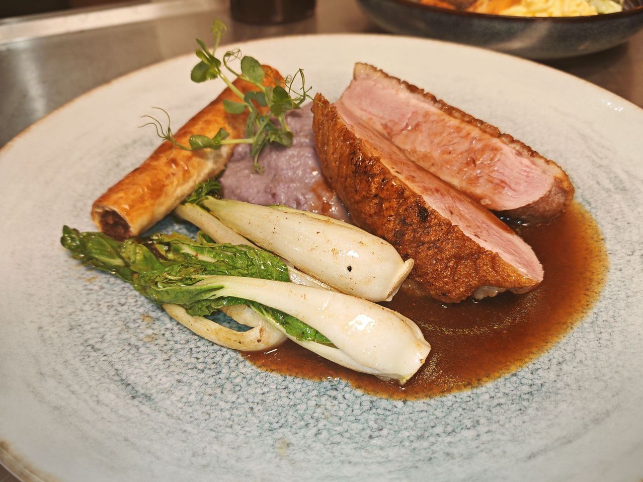 Ein Teller mit Gourmetgericht, bestehend aus gebratenem Schweinefleisch, lila Kartoffelpüree und grünem Gemüse mit brauner Sauce.