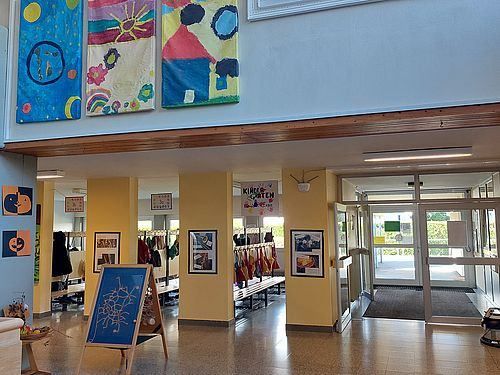 Das Innere eines Gebäudes mit gelben Wänden, das verschiedene farbenfrohe Kunstwerke und Bilder an den Wänden zeigt. Ein blaues Banner mit einem Design steht auf dem Boden. Eine Bank ist in der Nähe der Wand mit einem Schild platziert.