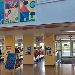 Ein Innenansicht eines Flurs mit gelben Wänden, der farbenfrohe Kinderkunstwerke, einen hölzernen Staffelei mit einer blauen Zeichnung und eine Glastür, die zu einem anderen Raum führt, zeigt.