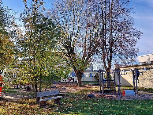 Ein Spielplatz mit Herbstlaub, bestehend aus einer Holzbank, Bäumen und einer Kletterstruktur. Im Hintergrund befindet sich ein Gebäude mit blauen Fensterläden.