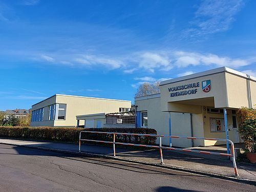 Das Volksschule Krensdorf Schulgebäude steht vor einem blauen Himmel mit Wolken im Hintergrund. Das Gebäude hat ein modernes Design mit Glasfenstern und einem Logo an der Vorderseite.