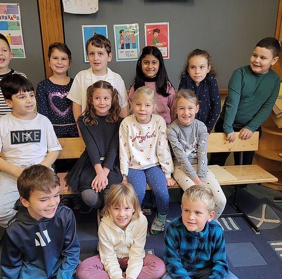 Eine Gruppe von Kindern in einem Klassenzimmer, die auf Bänken sitzen und für ein Foto lächeln. Hinter ihnen hängen Poster an der Wand und rechts sind Holzregale.