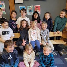 Eine Gruppe von Kindern in einem Klassenzimmer, die auf Bänken sitzen und für ein Foto lächeln. Hinter ihnen hängen Poster an der Wand und rechts sind Holzregale.