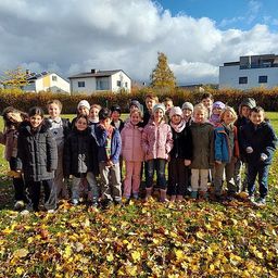Eine Gruppe von Kindern in Winterkleidung steht auf einem Feld aus gefallenen Blättern, mit Häusern und Grünpflanzen im Hintergrund.