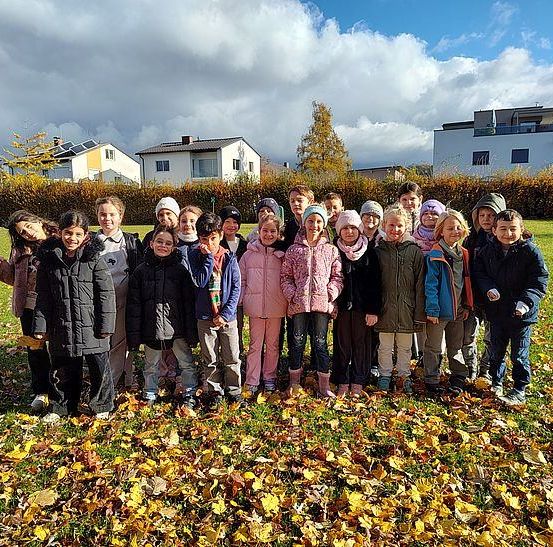 Eine Gruppe von Kindern in Winterkleidung steht auf einem Feld aus gefallenen Blättern, mit Häusern und Grünpflanzen im Hintergrund.