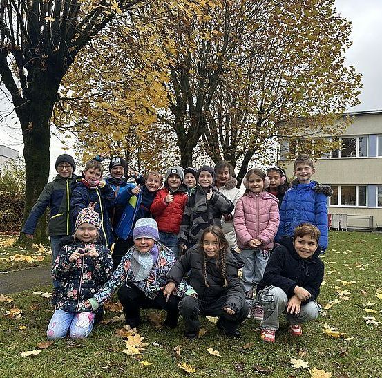 Eine Gruppe von Kindern posiert für ein Foto auf einer Rasenfläche mit Bäumen und einem Gebäude im Hintergrund.
