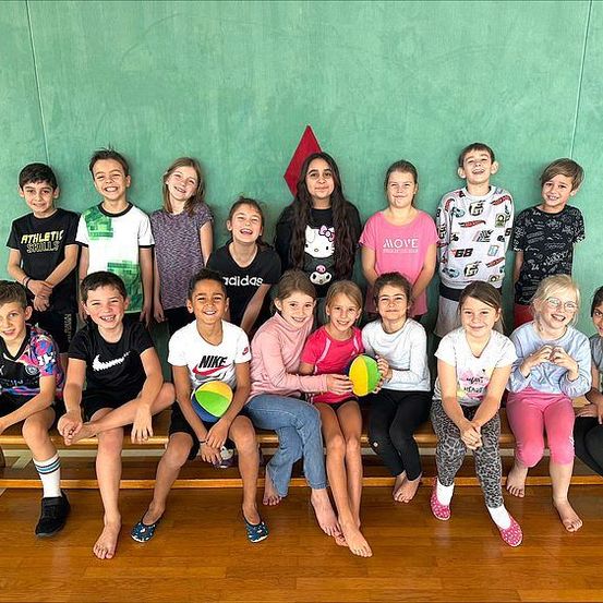 Eine Gruppe junger Kinder sitzt auf einer Holzbank in einer Turnhalle und lächelt für ein Foto mit grünen Wänden im Hintergrund.