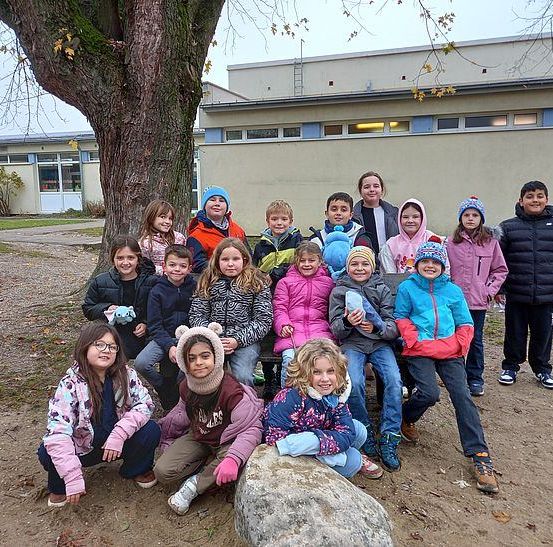 Eine Gruppe von Kindern und zwei Erwachsenen posiert für ein Foto im Freien, mit einem Baum und einem Gebäude im Hintergrund.