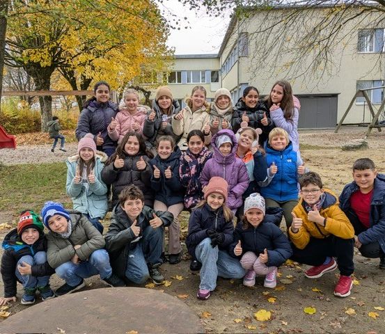 Eine Gruppe junger Schüler posiert für ein Foto im Freien. Sie alle lächeln und geben Daumen hoch. Der Boden ist mit trockenen Blättern bedeckt. Hinter ihnen steht ein Gebäude mit Glasfenstern.