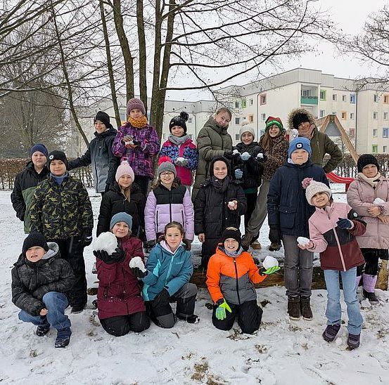 Eine Gruppe von Kindern in Winterkleidung posiert für ein Foto im Schnee, mit Bäumen und Gebäuden im Hintergrund.
