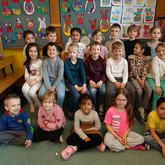 Eine Gruppe junger Kinder sitzt zusammen in einem Klassenzimmer. Einige sitzen auf dem Boden, während andere auf einer Holzbank sitzen. Osterdekorationen hängen an der Wand hinter ihnen.