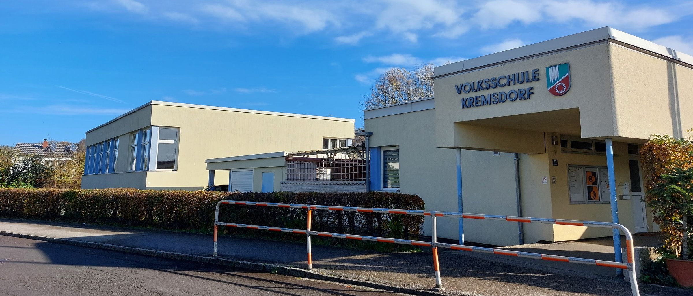 Ein sonniger Tag mit verstreuten Wolken, die Vorderansicht eines Schulgebäudes mit dem Schild Volksschule Kremsdorf über dem Eingang. Es gibt zwei Gebäude, eines mit Balkon und das andere mit einem Tor.