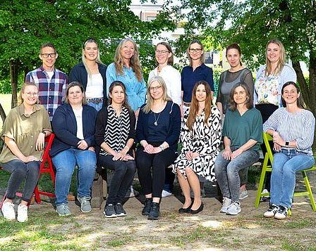 Eine Gruppe von Menschen sitzt auf verschieden farbigen Stühlen in einem Park mit Bäumen im Hintergrund. Sie alle lächeln und posieren für ein Foto.