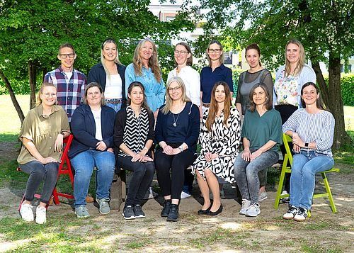 Eine Gruppe von Menschen sitzt auf verschieden farbigen Stühlen in einem Park mit Bäumen im Hintergrund. Sie alle lächeln und posieren für ein Foto.