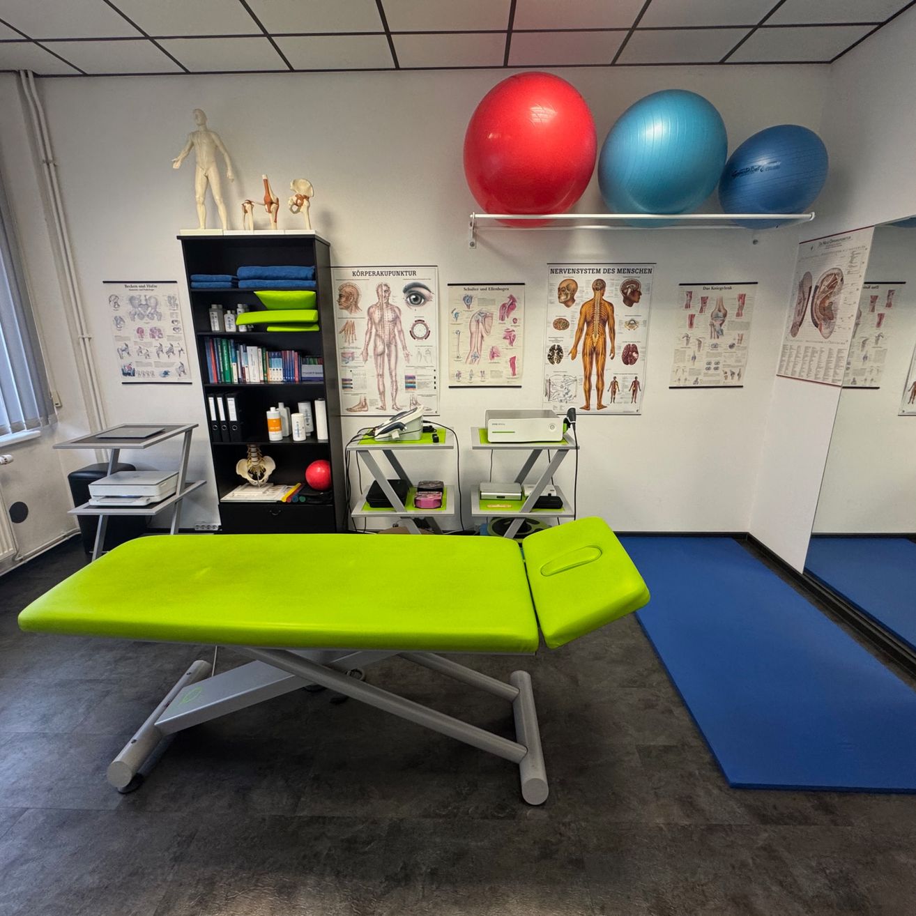 Ein Behandlungsraum mit einem grünen Untersuchungstisch, drei großen Gymnastikbällen, einem Bücherregal und anatomischen Postern an der Wand. Es gibt auch zwei blaue Gymnastikmatten auf dem Boden.