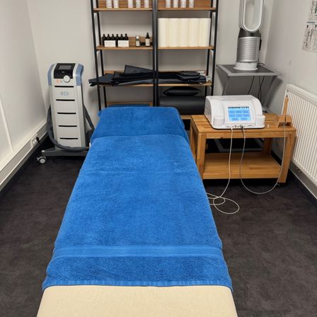 Ein Raum mit einem blauen Massagetisch, der mit einem blauen Handtuch bedeckt ist. Ein medizinisches Gerät steht auf einem Holztisch neben dem Tisch. Regale mit Flaschen und Handtüchern stehen an der Wand.