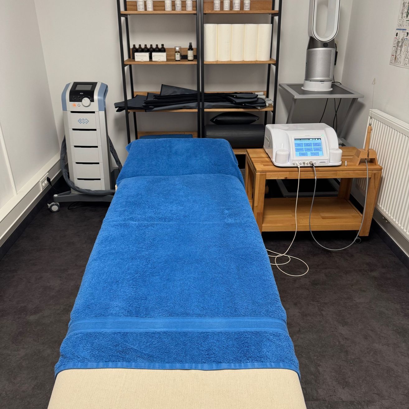 Ein Raum mit einem blauen Massagetisch, der mit einem blauen Handtuch bedeckt ist. Ein medizinisches Gerät steht auf einem Holztisch neben dem Tisch. Regale mit Flaschen und Handtüchern stehen an der Wand.