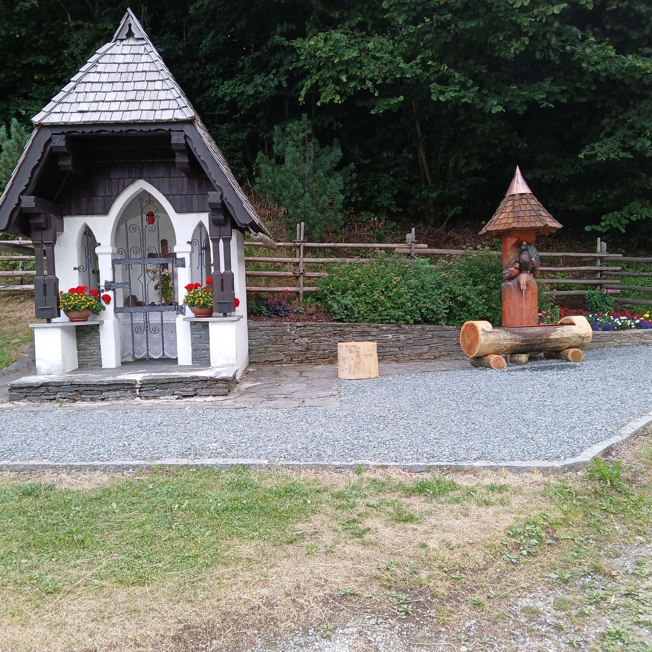 Eine kleine weiße Kapelle mit einem spitzen Dach steht in einem Kiesbereich, geschmückt mit roten Blumen. Ein steinerner Brunnen ist in der Nähe, und ein Holzzaun begrenzt das Gebiet.