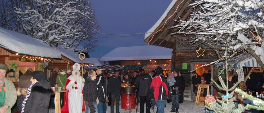 Menschen versammeln sich in einem schneebedeckten Außenbereich für eine Veranstaltung, mit einer Santa Claus-Figur, festlichen Lichtern und einem kleinen Tisch mit Tassen. Holzhütten und Bäume sind in der Umgebung, und ein kleiner roter Fass steht in der Mitte.