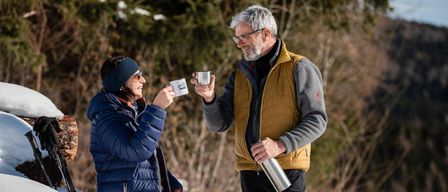 Ein Mann und eine Frau sitzen auf einem Schneehaufen, lächeln und halten Edelstahlbecher. Die Frau trägt eine Mütze und eine blaue Jacke, und der Mann trägt eine gelbe Weste und eine Brille. Sie halten beide Edelstahlbecher und der Mann einen Flakon.