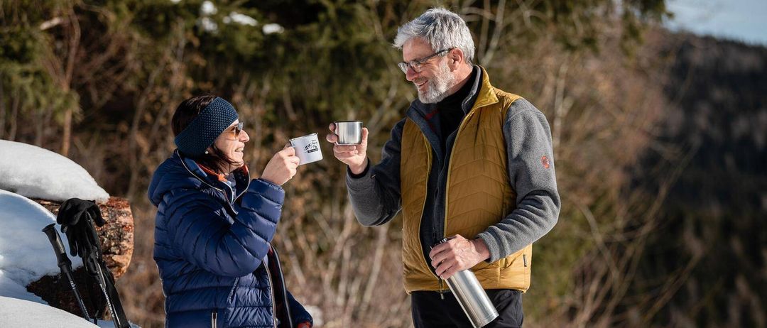 Ein Mann und eine Frau sitzen auf einem Schneehaufen, lächeln und halten Edelstahlbecher. Die Frau trägt eine Mütze und eine blaue Jacke, und der Mann trägt eine gelbe Weste und eine Brille. Sie halten beide Edelstahlbecher und der Mann einen Flakon.