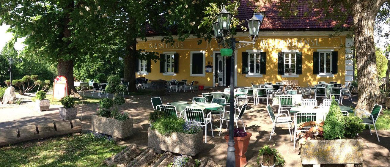 Ein Außenbereich mit vielen Tischen und Stühlen steht vor einem gelben Gebäude mit dem Namen "Gasthaus". Bäume und Topfpflanzen umgeben den Bereich.