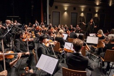 Eine Gruppe von Musikern spielt auf der Bühne, eine Frau steht vor ihnen und dirigiert wahrscheinlich.