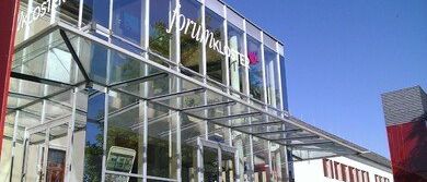 Ein modernes Gebäude mit Glaswänden und einem Glasdach, mit einem Schild, das "Brunklaus" und eine rote Wand auf der linken Seite zeigt.