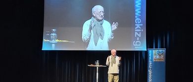 Ein älterer Mann steht auf der Bühne und hält eine Präsentation für ein Publikum. Ein großer Bildschirm hinter ihm zeigt ein Bild eines Mannes, und Banner mit Website-URLs sind an den Seiten der Bühne sichtbar.