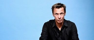 Ein Mann sitzt auf einem orangefarbenen Stuhl mit zusammengeklappten Händen, blickt nach vorne und posiert wahrscheinlich für ein Foto.