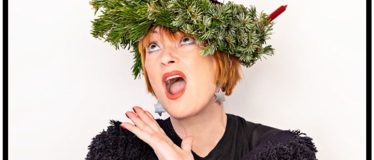 Eine Frau mit kurzen roten Haaren, die einen schwarzen Pullover und eine Weihnachtskrone aus Tannenzweigen trägt. Die Krone hat rote Kerzen.