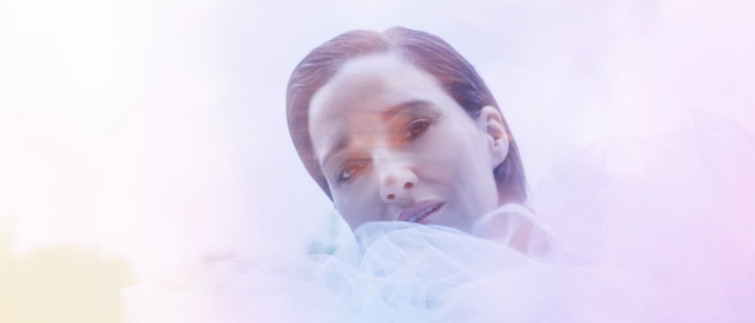 Eine Frau liegt auf der Seite mit einem verträumten, verschwommenen Effekt um sie herum, trägt ein weißes Tuch oder eine Schleier. Ihr Gesicht ist mit weichem Licht beleuchtet.