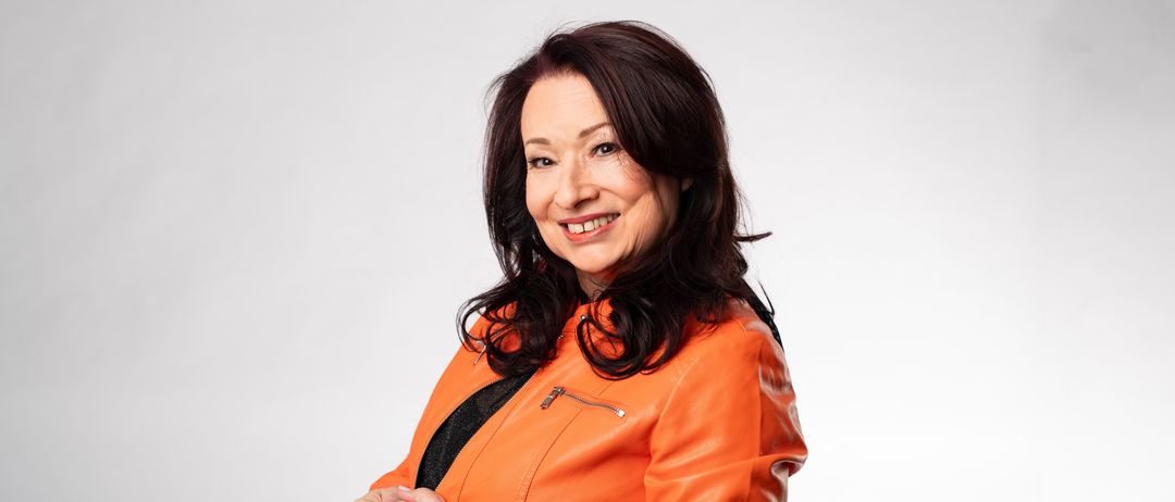 Eine Frau mit schwarzen Haaren, die eine orangefarbene Lederjacke trägt, steht lächelnd mit verschränkten Händen da.