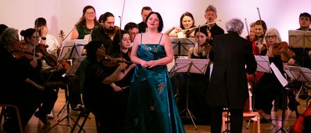 Eine Frau in einem blauen Kleid steht auf der Bühne vor einem Symphonieorchester. Die Musiker spielen Violinen und andere Instrumente, während der Dirigent vor ihnen steht.