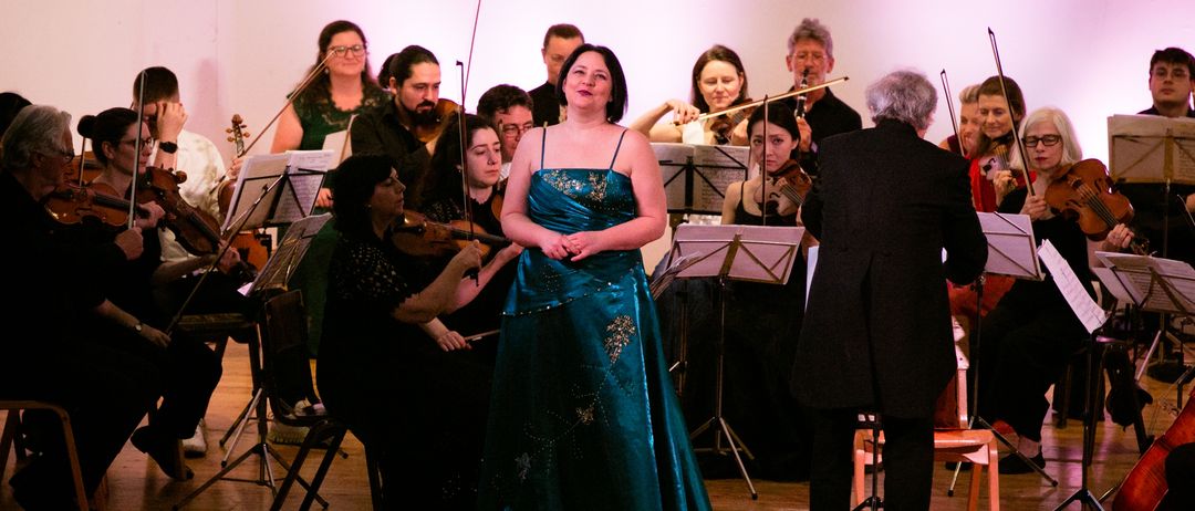 Eine Frau in einem blauen Kleid steht auf der Bühne vor einem Symphonieorchester. Die Musiker spielen Violinen und andere Instrumente, während der Dirigent vor ihnen steht.