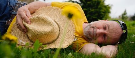 Ein Mann liegt auf dem Gras, trägt ein gelbes Shirt, einen Strohhut und eine Sonnenbrille. Er scheint zu ruhen, mit geschlossenen Augen.