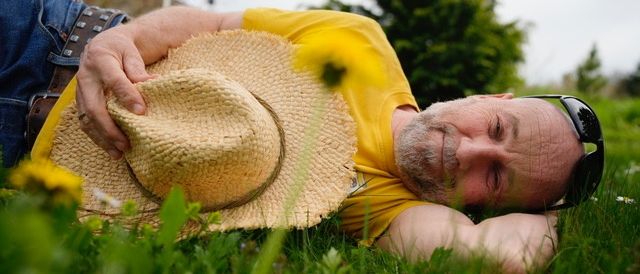 Ein Mann liegt auf dem Gras, trägt ein gelbes Shirt, einen Strohhut und eine Sonnenbrille. Er scheint zu ruhen, mit geschlossenen Augen.