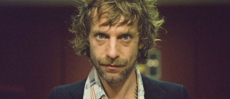 Ein Mann mit lockigem Haar und Bart steht in einem Raum, trägt ein kariertes Hemd und eine Halskette. Er schaut direkt in die Kamera.