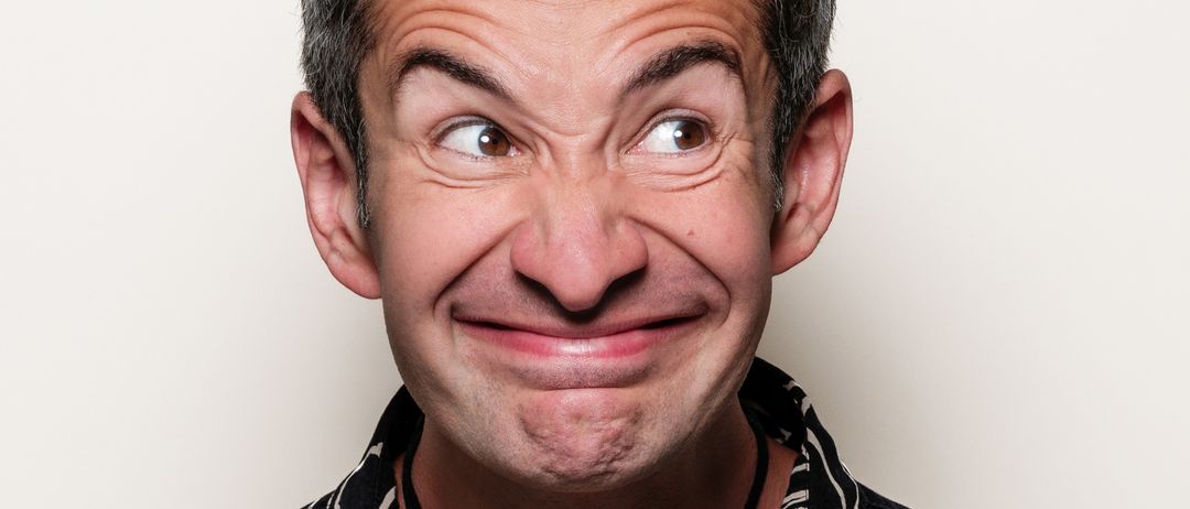 Ein Mann mit einem schelmischen Lächeln trägt ein schwarzes Blumenhemd und eine braune Halskette. Der Mann hat kurze Haare und steht vor einem schlichten weißen Hintergrund.