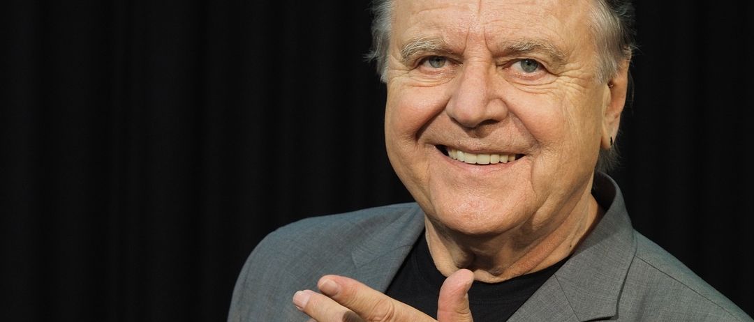 Ein älterer Mann mit grauem Haar und einem grauen Anzug lächelt und gibt einen Daumen hoch. Er trägt ein schwarzes Hemd und eine silberne Armbanduhr.