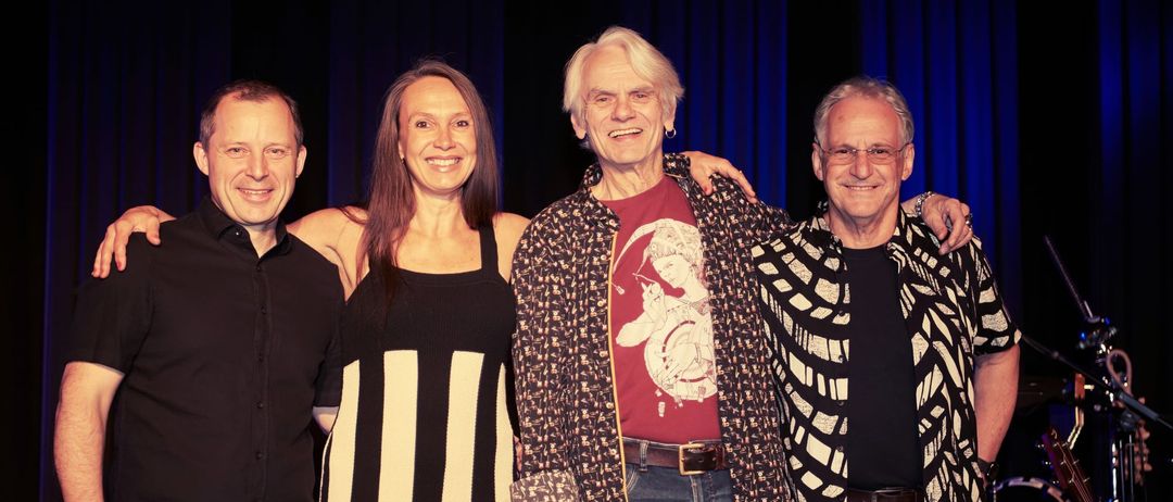 Vier Personen posieren für ein Foto auf einer Bühne mit blauen Vorhängen. Sie lächeln.