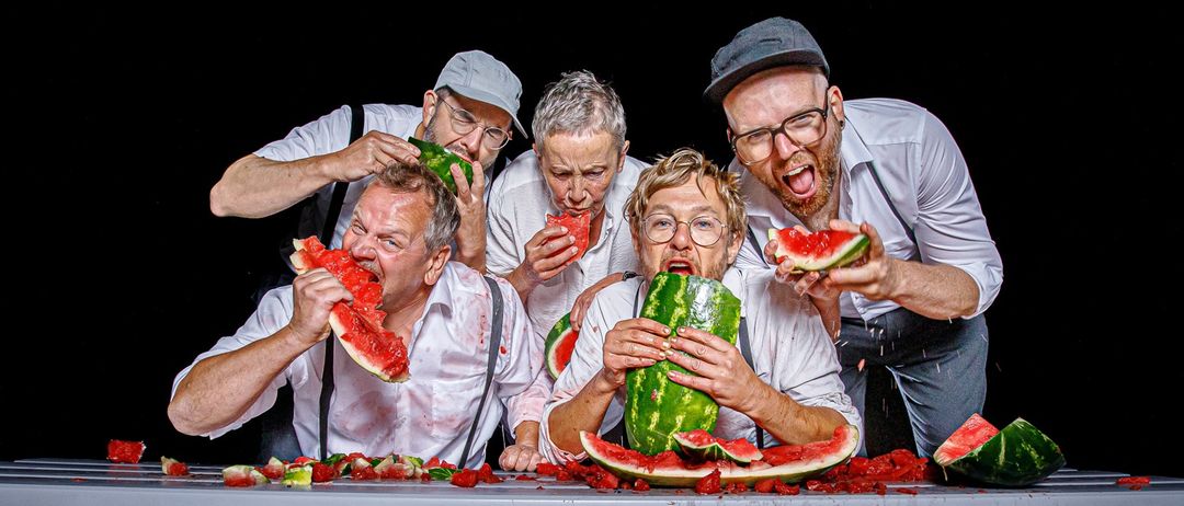 Fünf Männer mit Brillen und Hosenträger tragen an Wassermelonen auf einem Tisch und machen einen Schlamassel.