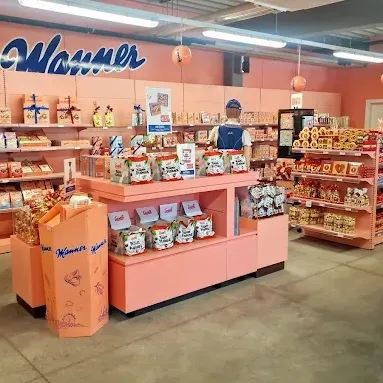 Ein Geschäft mit rosa Regalen präsentiert verschiedene Snacks und Produkte. Die Regale sind mit Kartons und Tüten gefüllt. Eine Person steht hinter einem Tresen, und ein Trolley befindet sich in der Nähe.