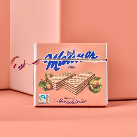 Eine rosafarbene Box von 'Munzer' Waffeln mit einem Fairtrade-Kakao-Label, platziert auf einem rosa Hintergrund. Die Box ist teilweise geöffnet und zeigt zwei gestapelte Waffeln.