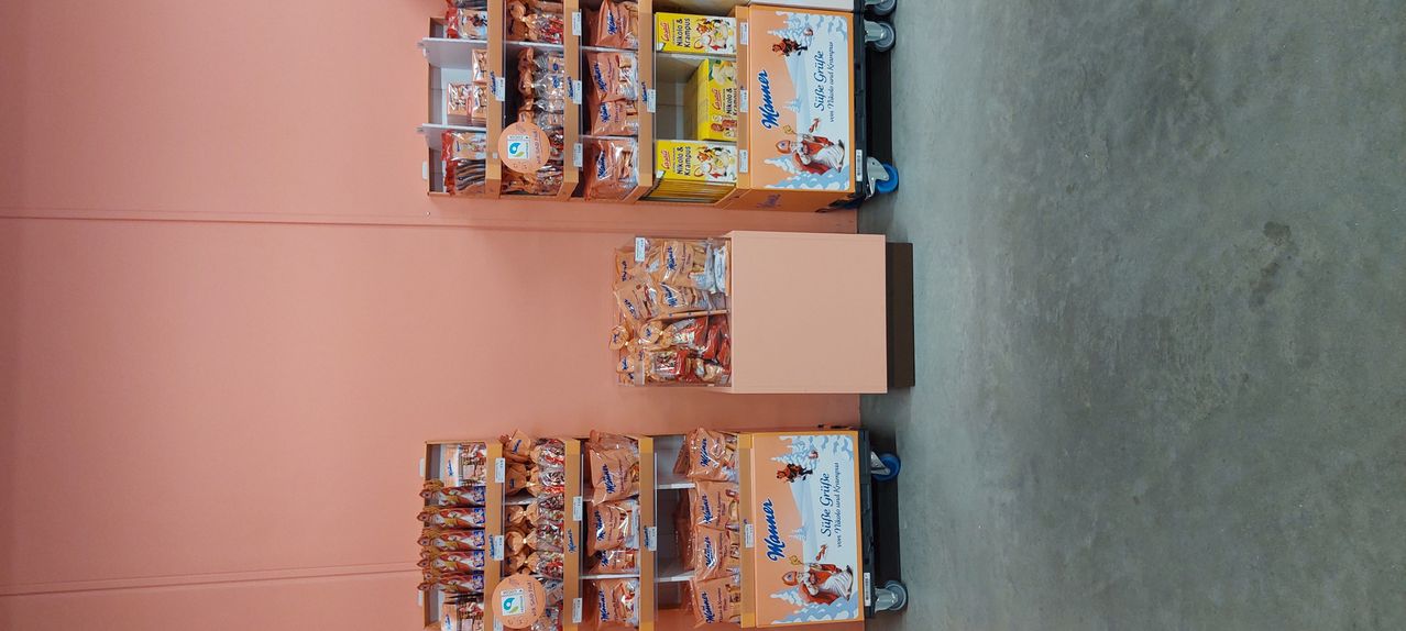 Das Bild zeigt einen Laden mit verschiedenen Snack-Displays auf Regalen. Die Regale sind mit Tüten verschiedener Snacks gefüllt. Einige der Snacks haben ein Weihnachtsthema. Die Regale sind in Reihen angeordnet. Jede Reihe hat eine Schachtel mit Weihnachtsmann-Snacks. Der Laden hat eine rosa Wand.