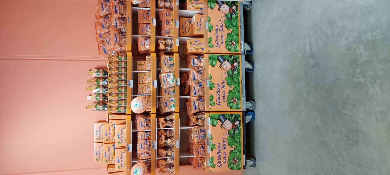 Ein Supermarktregal zeigt Schachteln von 'Glückliches Neues Jahr', einem deutschen Neujahrs-Feierprodukt. Die Schachteln sind in Orange und Grün, mit einem Cartoon-Charakter und ordentlich auf rollenden Regalen gestapelt.